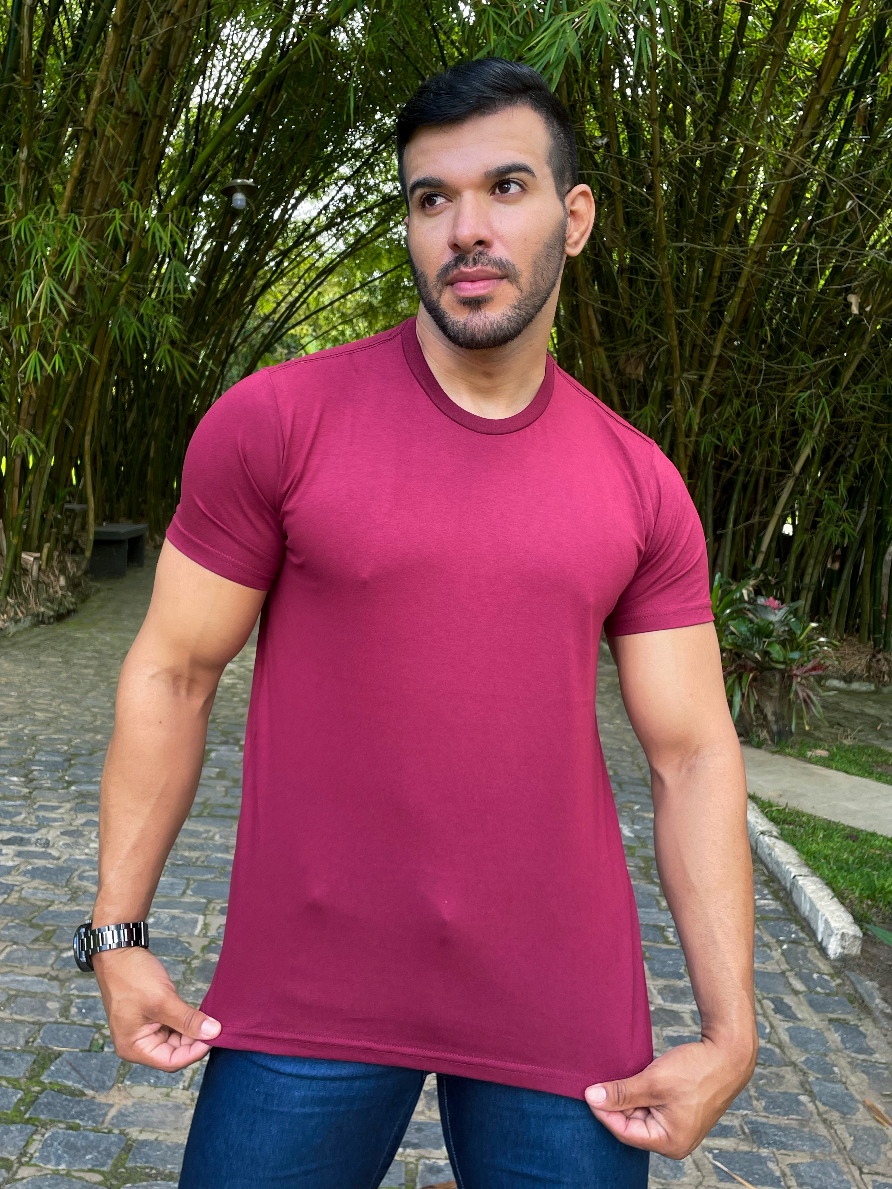 Camiseta Vinho Homem posando ao ar livre, vestindo uma camiseta vinho e jeans, com um fundo de bambus verdes.